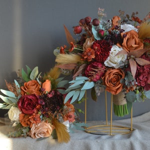 May include: Two bouquets of artificial flowers in shades of orange, red, and white. The larger bouquet is on a gold stand and the smaller bouquet is on a beige surface.