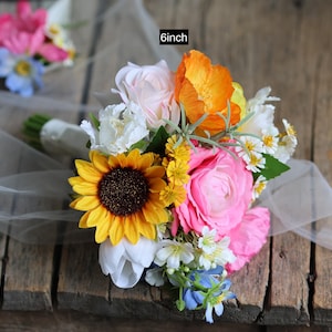May include: A floral bouquet featuring a large yellow sunflower, bright pink ranunculus, orange poppies, white tulips, and small blue and yellow filler flowers. The bouquet is wrapped in white ribbon and measures 6 inches in diameter.