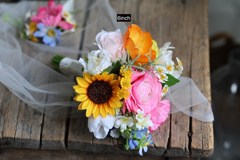 May include: A floral bouquet featuring a large yellow sunflower, bright pink ranunculus, orange poppies, white tulips, and small blue and yellow filler flowers. The bouquet is wrapped in white ribbon and measures 6 inches in diameter.