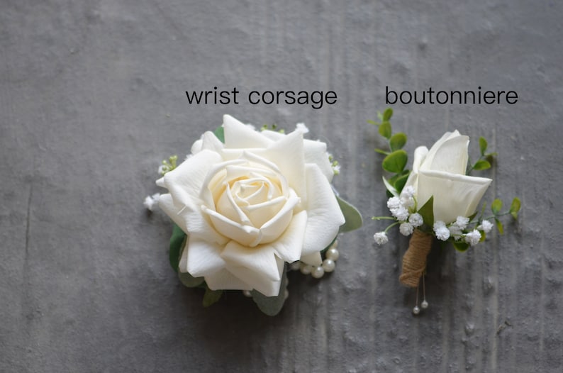 Puede incluir: Dos boutonni&egrave;res de rosas blancas, una con una sola rosa y la otra con una rosa m&aacute;s grande y detalles de perlas. El texto "wrist corsage" y "boutonniere" est&aacute; escrito sobre los arreglos.