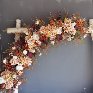Peut inclure: Une arche en bois rustique décorée d'une guirlande de fleurs artificielles dans des tons d'orange, de brun et de crème. Les fleurs sont disposées en cascade, créant un look romantique et élégant.