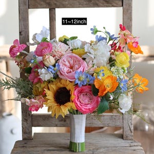 May include: A vibrant floral bouquet featuring a mix of pink, yellow, orange, white, and blue artificial flowers, including a prominent sunflower and roses. The stems are wrapped in white tape, and the bouquet measures 11 to 12 inches in width.