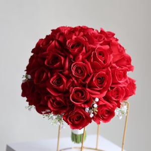 Puede incluir: Un ramo redondo de rosas rojas vibrantes, acentuado con pequeñas flores blancas y vegetación. El ramo se sujeta con una cinta verde y se presenta sobre un soporte dorado, creando un arreglo floral clásico y elegante.