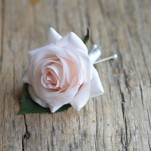 Pode incluir: Uma única rosa rosa claro com folhas verdes, presa a um alfinete prateado. A rosa está sobre uma superfície de madeira rústica.