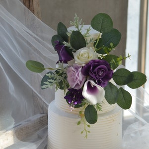 Può includere: Una torta nuziale bianca a due piani decorata con un'arrangiamento floreale viola e bianco. L'arrangiamento include calle, rose e verde.