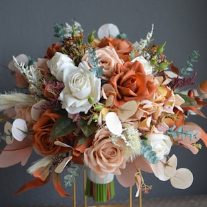 Puede incluir: Un ramo de flores artificiales en tonos melocotón, blanco y naranja quemado. El ramo incluye rosas, eucalipto y otras plantas verdes.