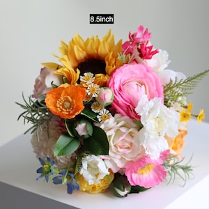 May include: A floral bouquet measuring 8.5 inches in diameter, featuring a vibrant mix of yellow sunflowers, bright pink ranunculus, orange poppies, white roses, and small blue flowers, accented with green foliage and various small filler blossoms.