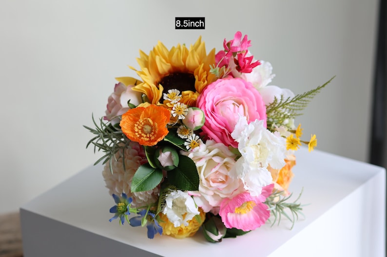 May include: A floral bouquet measuring 8.5 inches in diameter, featuring a vibrant mix of yellow sunflowers, bright pink ranunculus, orange poppies, white roses, and small blue flowers, accented with green foliage and various small filler blossoms.