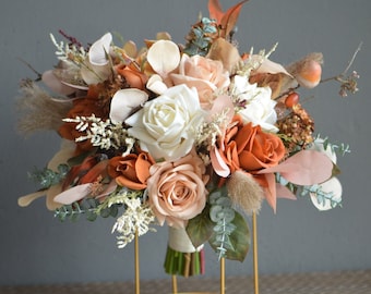 May include: A bouquet of faux flowers in shades of peach, white, and burnt orange. The bouquet includes roses, eucalyptus, and other greenery. The bouquet is arranged in a gold metal stand.