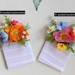 May include: Two pocket boutonnieres labeled #1 and #2. Each features a white rectangular fabric base topped with artificial flowers. #2 includes orange, blue, and pink blooms with green foliage. #1 features pink, blue, and yellow flowers with greenery.