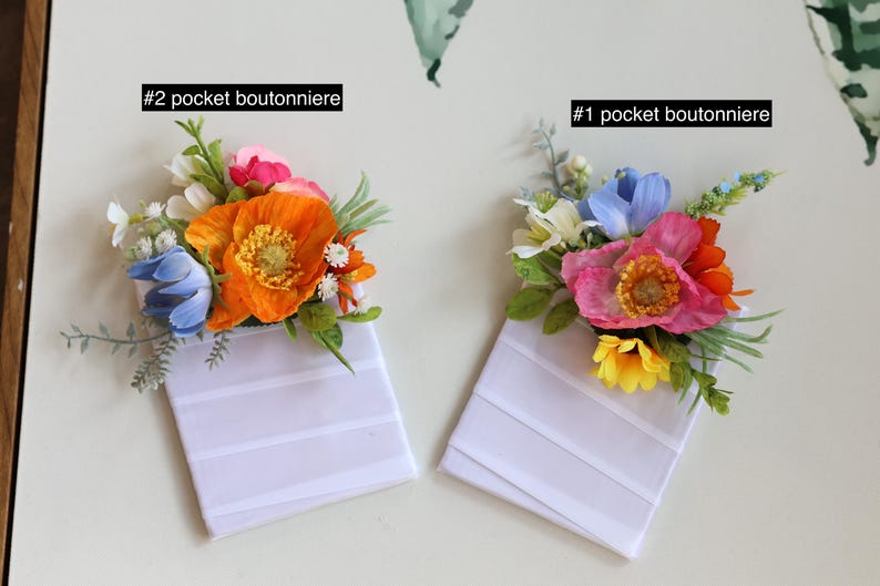 May include: Two pocket boutonnieres labeled #1 and #2. Each features a white rectangular fabric base topped with artificial flowers. #2 includes orange, blue, and pink blooms with green foliage. #1 features pink, blue, and yellow flowers with greenery.