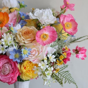 May include: A vibrant floral arrangement featuring a mix of orange, pink, yellow, white, and blue flowers. The bouquet includes roses, poppies, and small white blossoms with green foliage, all gathered together in a white vase against a plain background.