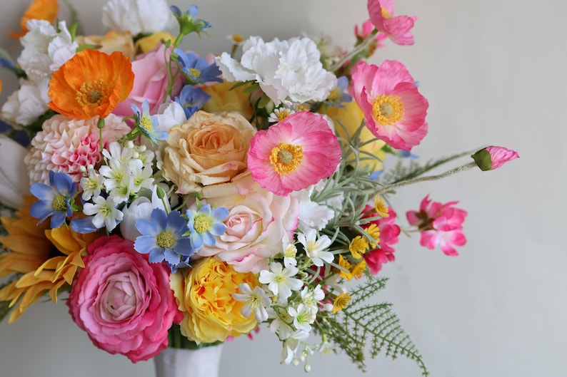 May include: A vibrant floral arrangement featuring a mix of orange, pink, yellow, white, and blue flowers. The bouquet includes roses, poppies, and small white blossoms with green foliage, all gathered together in a white vase against a plain background.