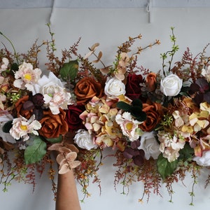 May include: A garland of artificial flowers in shades of burgundy, white, and orange. The garland is made up of roses, hydrangeas, and other small flowers. It is perfect for adding a touch of fall color to your home decor.