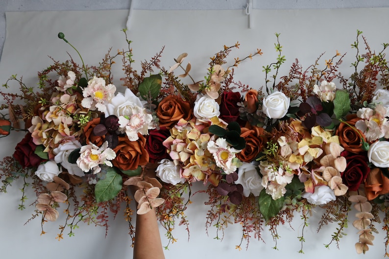May include: A garland of artificial flowers in shades of burgundy, white, and orange. The garland is made up of roses, hydrangeas, and other small flowers. It is perfect for adding a touch of fall color to your home decor.