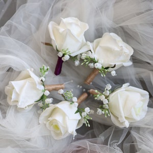 Könnte beinhalten: Fünf weiße Rosen-Ansteckblumen mit kleinen weißen Blüten und grünen Blättern. Die Ansteckblumen sind in einem Kreis auf einem weißen Tüllstoff angeordnet.