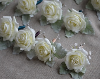 May include: A collection of white rose boutonnieres with greenery and small white flowers. The boutonnieres are arranged in a semi-circle on a textured fabric surface.