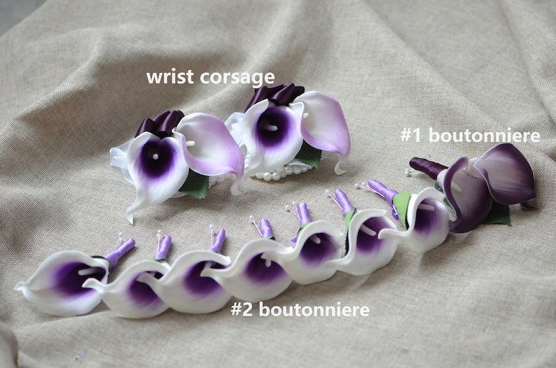 May include: A set of purple and white calla lily boutonnieres and a wrist corsage. The boutonnieres have purple ribbon and the corsage has a white ribbon.