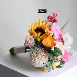 May include: A floral bouquet featuring a large yellow sunflower, pink roses, orange poppies, white carnations, and small blue flowers with green foliage. The bouquet has a wrapped stem and a label indicating a height of 8.5inch.