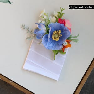 May include: A pocket boutonniere labeled #3 pocket boutonniere featuring a cluster of artificial flowers including a large blue poppy, smaller white and orange blossoms, and green foliage attached to a white rectangular fabric pocket square base.