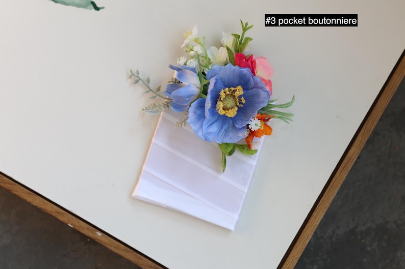 May include: A pocket boutonniere labeled #3 pocket boutonniere featuring a cluster of artificial flowers including a large blue poppy, smaller white and orange blossoms, and green foliage attached to a white rectangular fabric pocket square base.