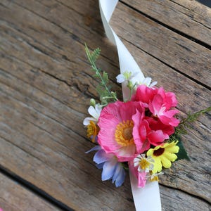 May include: A floral wrist corsage featuring a cluster of artificial pink poppies, yellow daisies, and small white flowers with green foliage. The arrangement is attached to a long, smooth white satin ribbon that extends from the floral center.