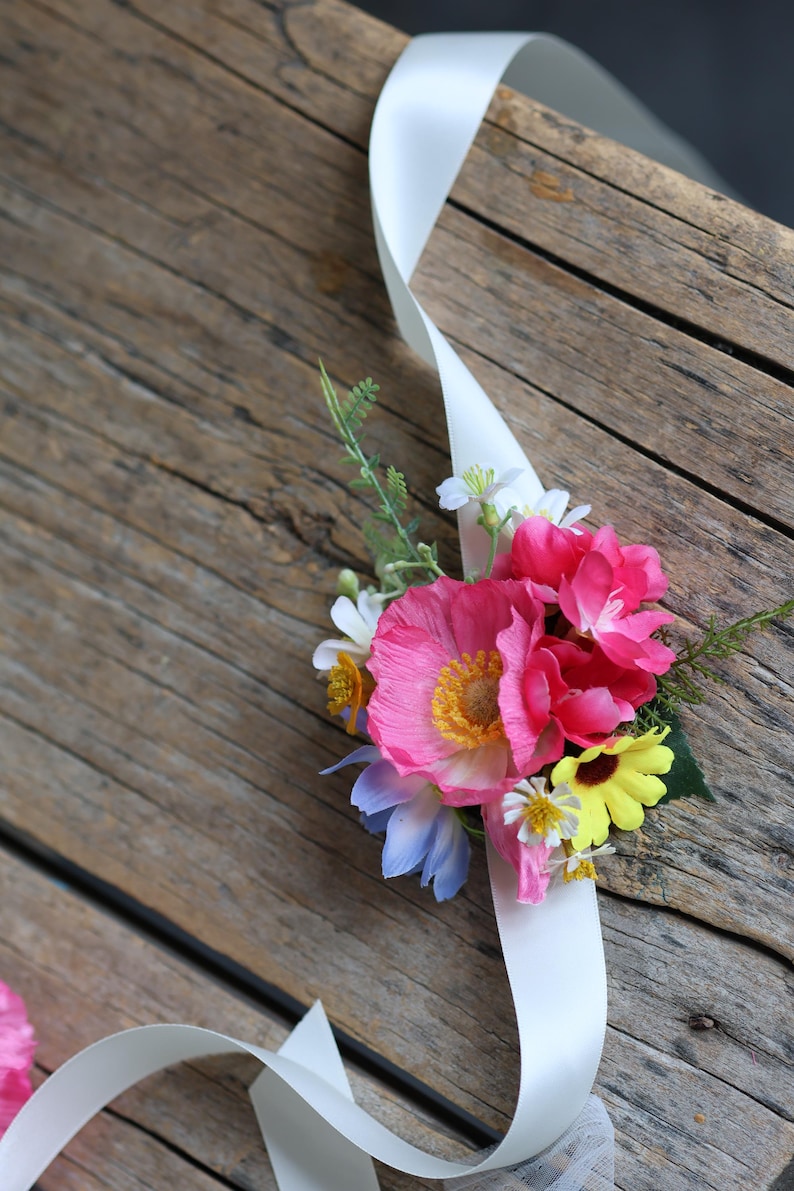 May include: A floral wrist corsage featuring a cluster of artificial pink poppies, yellow daisies, and small white flowers with green foliage. The arrangement is attached to a long, smooth white satin ribbon that extends from the floral center.