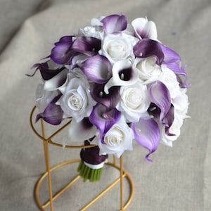 May include: A bridal bouquet made of white roses and purple calla lilies. The bouquet is arranged in a round shape and is held together with a purple ribbon.