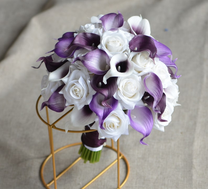 May include: A bridal bouquet made of white roses and purple calla lilies. The bouquet is arranged in a round shape and is held together with a purple ribbon.