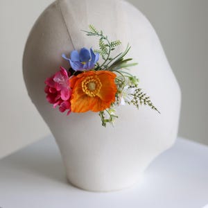 May include: A floral hair accessory featuring a vibrant orange poppy, a bright pink flower, a small blue blossom, and green foliage. The arrangement is attached to a thin, neutral-colored band, creating a colorful botanical hair piece.