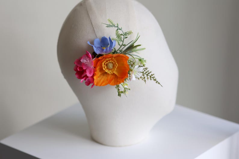 May include: A floral hair accessory featuring a vibrant orange poppy, a bright pink flower, a small blue blossom, and green foliage. The arrangement is attached to a thin, neutral-colored band, creating a colorful botanical hair piece.