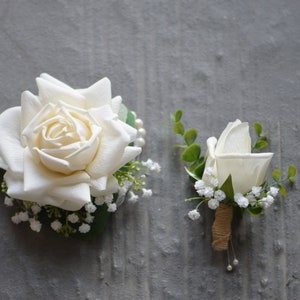 May include: Two white rose boutonnieres with greenery and small white flowers. The larger boutonniere has a single white rose with a pearl accent. The smaller boutonniere has a single white rose with a brown twine accent.