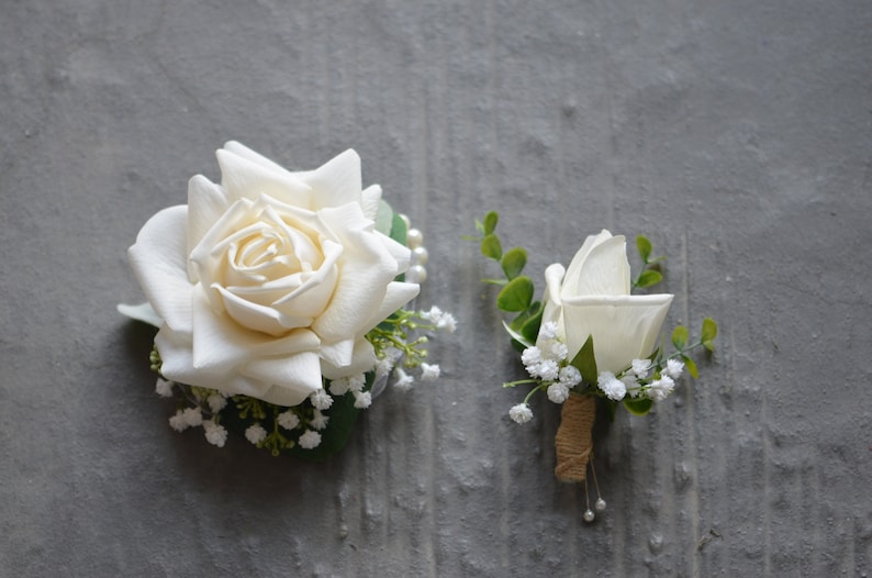 Puede incluir: Dos boutonni&egrave;res de rosas blancas con vegetaci&oacute;n y peque&ntilde;as flores blancas. La boutonni&egrave;re m&aacute;s grande tiene una sola rosa blanca con un acento de perla. La boutonni&egrave;re m&aacute;s peque&ntilde;a tiene una sola rosa blanca con un acento de cuerda marr&oacute;n.