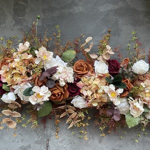 May include: A garland of artificial flowers in shades of burgundy, orange, and cream. The garland features roses, hydrangeas, and other floral elements.