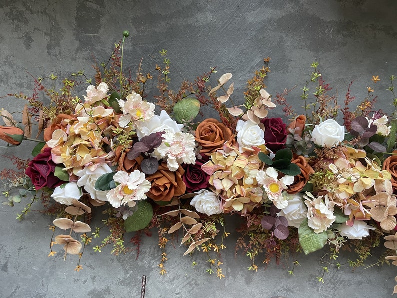 May include: A garland of artificial flowers in shades of burgundy, orange, and cream. The garland features roses, hydrangeas, and other floral elements.
