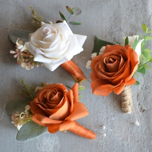 Puede incluir: Tres boutonnières con rosas naranjas y blancas y vegetación. Las boutonnières están atadas con cuerda y tienen alfileres de perla.