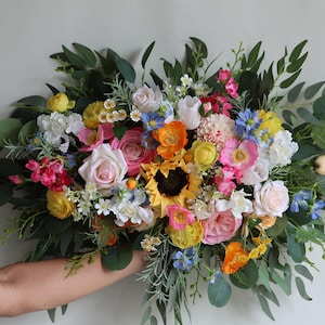 May include: A large, horizontal floral arrangement featuring a vibrant mix of sunflowers, pink roses, orange poppies, blue cornflowers, and white blossoms, accented with abundant green eucalyptus leaves and various sprigs of filler greenery.