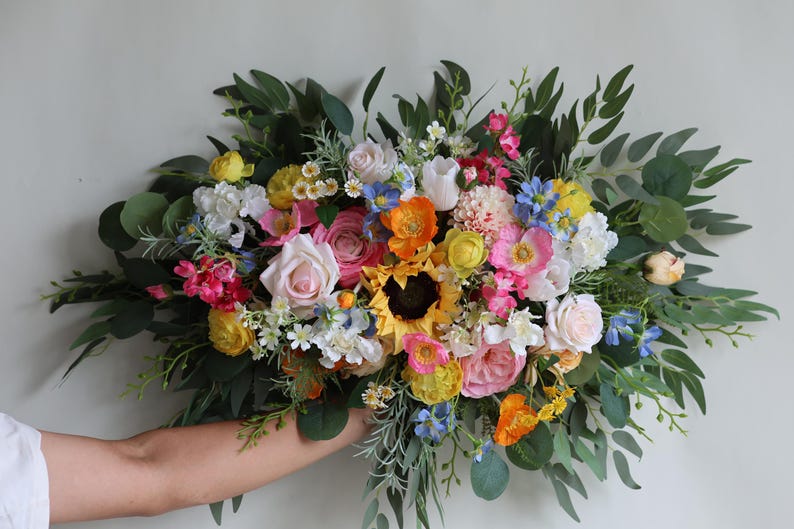 May include: A large, horizontal floral arrangement featuring a vibrant mix of sunflowers, pink roses, orange poppies, blue cornflowers, and white blossoms, accented with abundant green eucalyptus leaves and various sprigs of filler greenery.