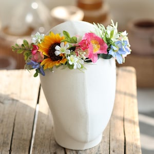 May include: A floral crown featuring a vibrant mix of artificial flowers including a large yellow sunflower, bright pink blossoms, small white flowers, and light blue petals, accented with green leaves and foliage, displayed on a white head form.