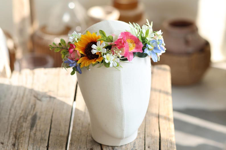 May include: A floral crown featuring a vibrant mix of artificial flowers including a large yellow sunflower, bright pink blossoms, small white flowers, and light blue petals, accented with green leaves and foliage, displayed on a white head form.