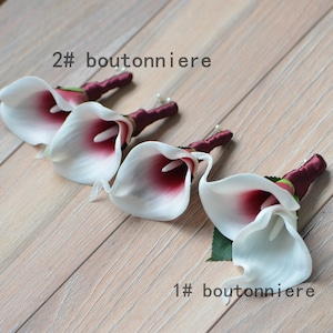 May include: Three white calla lily boutonnieres with burgundy accents and burgundy ribbon. The boutonnieres are arranged in a row on a wooden surface. The text "2# boutonniere" is above the boutonnieres and the text "1# boutonniere" is below the boutonnieres.