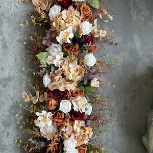 May include: A long garland of artificial flowers with a mix of white, burgundy, and orange roses, hydrangeas, and greenery.