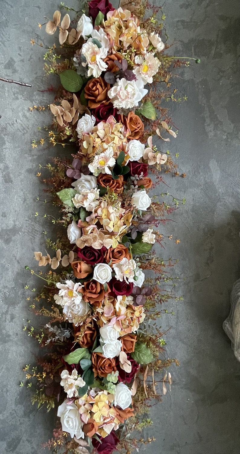 May include: A long garland of artificial flowers with a mix of white, burgundy, and orange roses, hydrangeas, and greenery.