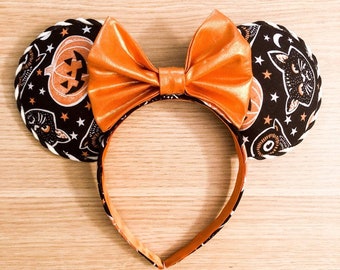 May include: Halloween-themed headband with black ears patterned with pumpkins, cats, and stars. The headband features a large orange bow and a matching patterned band, perfect for festive occasions.