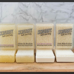 May include: Four clear plastic containers with natural soap infused scrubbers. The scrubbers are labeled with the scents: Olive Oil & Lavender, Olive Oil & Aloe, Goat Milk & Lavender, and Goat Milk & Aloe.  The scrubbers are sitting on a wooden surface.