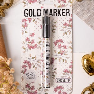 May include: A gold marker with a chisel tip, packaged on a white card with a floral design. The card reads "GOLD MARKER" in black text. The marker itself is decorated with a floral pattern and the words "GOLD MARKER".