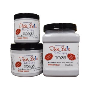 Op de afbeelding: Drie potten Dixie Belle Paint Company BOSS verf in grijs. De potten zijn gelabeld met de bedrijfsnaam, productnaam en maat. De kleinste pot is 8 ounce, de middelste pot is 16 ounce en de grootste pot is 32 ounce. De potten zijn allemaal gelabeld met "Problem solver!" en "Use only when needed!"