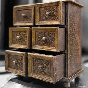 Könnte beinhalten: Ein kleiner Holzschrank mit sechs Schubladen, dunkelbraun lackiert und mit goldenen Verzierungen versehen. Jede Schublade hat ein Blumenmuster und einen kleinen, runden Knopf. Der Schrank steht auf kurzen, abgerundeten Füßen, und die Seitenwände haben ein sich wiederholendes Muster.