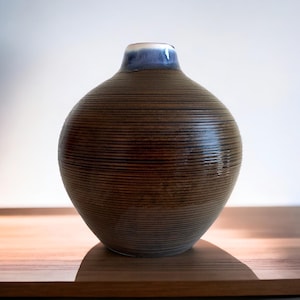 May include: A brown ceramic vase with horizontal textured lines. The vase has a rounded body and a narrow neck with a blue and white rim. The vase is sitting on a wooden surface.