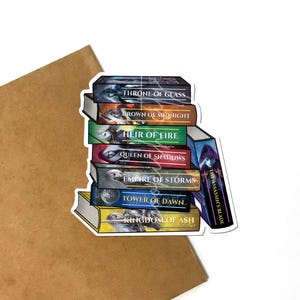 May include: A sticker featuring a stack of fantasy book covers with titles like "Throne of Glass" and "Kingdom of Ash." The books are in various colors, including blue, green, and yellow, with detailed illustrations. The sticker is on a white background.